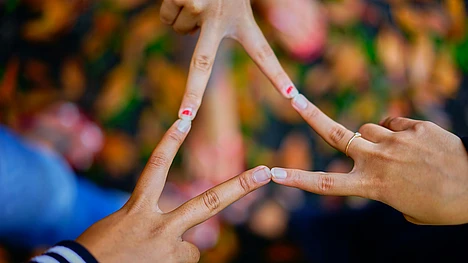 Four hands with fingers touching create a star shape against a blurred background of colorful leaves. One hand wears a ring, and another has nail art with red hearts.