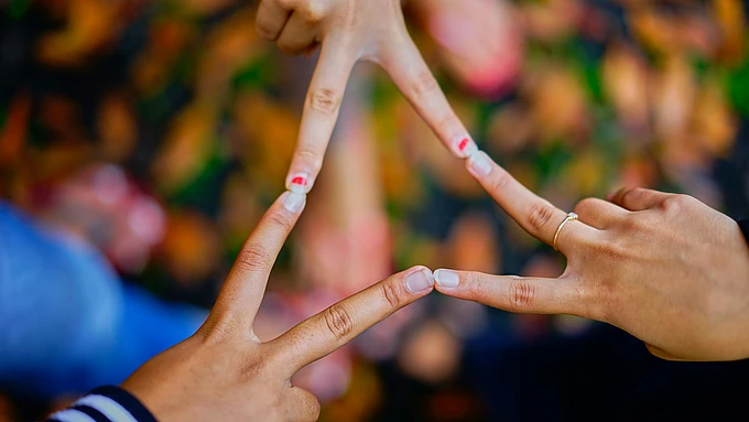 Four hands with fingers touching create a star shape against a blurred background of colorful leaves. One hand wears a ring, and another has nail art with red hearts.