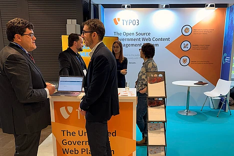 Four people talk at a TYPO3 booth with banners about open source government web content management. Two men stand in front, while two others stand near the booth’s display and a table with chairs is in the background.