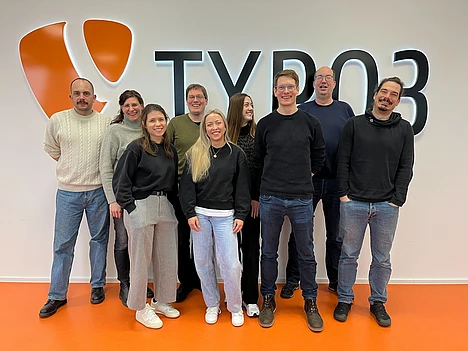 Nine people, standing and smiling in two rows, pose in front of a wall with a large TYPO3 logo. The floor is orange, and everyone appears relaxed and casual.
