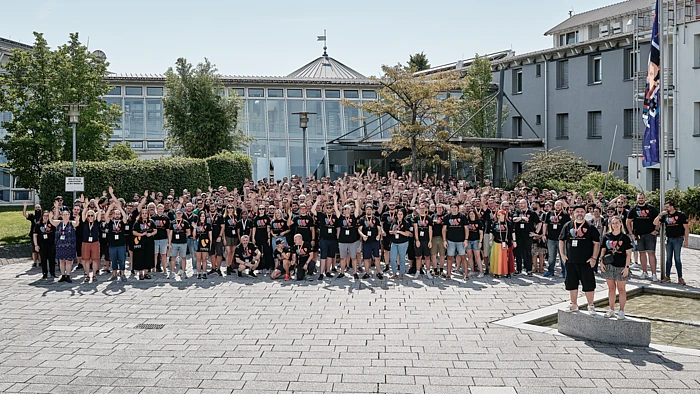 A large group of people wearing matching black T-shirts poses and cheers for a photo outside a modern building with glass windows on a sunny day. Trees and flags are visible in the background.