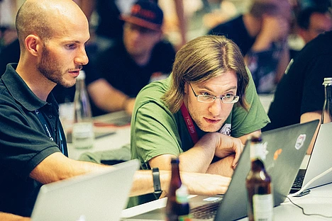 Two people sit at a table, intently looking at a laptop. One is pointing at the screen while the other focuses on it. Bottles and another laptop are on the table; other people are blurred in the background.